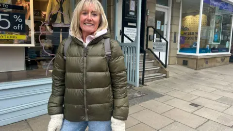 A woman with blonde hair and a dark green puffer coat, wearing cream mittens and smiling outside a shop in Ilkley.