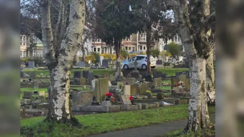 Ian Calway A graveyard dotted with silver birch trees. A car is visible in the background, having been squeezed on to one of the footpaths. 