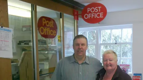 Contributed A man wearing a shirt standing next to a woman with short blonde hair. They are standing in front of a Post Office kiosk.