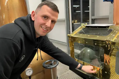 Ryan is leaning over to turn a handle on part of a machinery inside the distillery. 