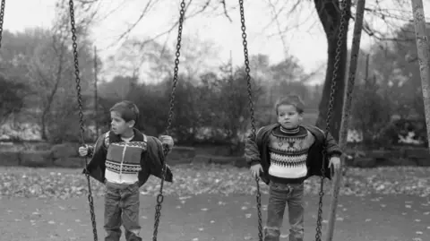 Sharon Hepburn Two young children stand on swings in a park, with trees and fallen leaves behind them.
