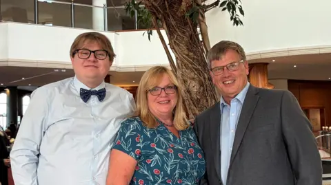 James Evans James Evans stands on the left with his parents who adopted him in 2004, Ruth and Andrew. James is wearing a pale blue shirt and a black bow-tie. He has blonde hair. Ruth is shorter than both James and Andrew and stands in the middle. She is wearing a teal dress with red berries and leaves as a pattern. She has blonde hair down to her shoulders. Andrew is stood next to her on the right wearing a light blue shirt and a grey suit jacket. He has short brown hair and is wearing glasses. 