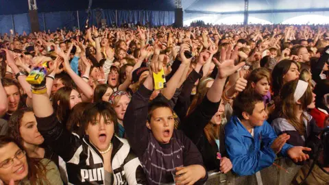 A crowd of young people cheer and dance with arms in the air in an enormous blue marquee. Several are clutching yellow disposable cameras.