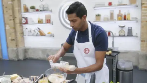 Shine TV/BBC Anurag Aggarwal wearing a white apron over a navy polo shirt while cooking on MasterChef.