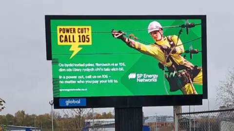 Padraig Durnin A large billboard advert for SP Energy Networks in London, showing an electricity worker working on wires, with wording in both English and Welsh saying: Os bydd toriad pwer, ffoniwch ni ar 105, dim ots i bwy rydych chi'n talu eich bil

In a power cut, call us on 105, no matter who you pay your bill to.