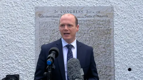 Gordon Lyons is standing in front of large print of the US Declaration Of Independence. He is wearing a navy suit and has two large microphones in front of him. 
