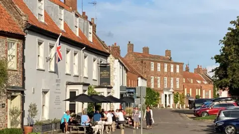 The view from the centre of Burnham Market. There are people siting at picnic tables outside a pub.