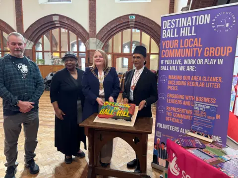 Pauling Black wears a black hat and jacket, and white shirt. She has two other women and a man to her right. On her left is a poster about Destination Ball Hill, Your local community group. She is holding a colourful cake with Welcome to Ball Hill iced on it.