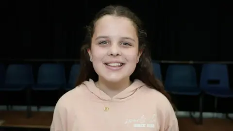 Qays Najm A girl with long brown hair stands wearing a pink hoody. She also has a gold chain necklace with small pedant.