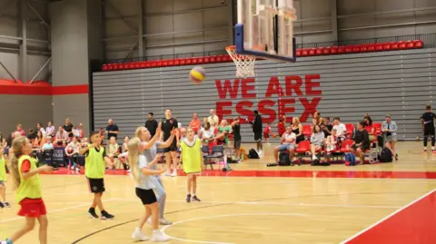 Children playing basketball at the Essex Sport Arena. There are six children on the court watching as a ball is thrown towards the net. A crowd of other youngsters and parents also watch on from the sideline.