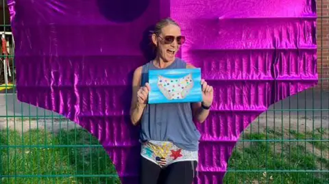 Charlotte Searle Charlotte grins while holding a drawing of a multi-coloured spotty knickers. She wears a blue strapped top and black leggings while wearing grey pants with red and blue stars. She satnds in front a huge decoration of a shiny purple pant pinned up on an outdoor railing.