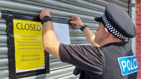 Cambridgeshire Police A police officer pinning paperwork on the metal shutters of the shop. There is a bright yellow order that says the shop is closed.