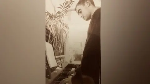 Supplied A brown and white sepier style photo of 'Bonehead' aka Paul Arthurs playing the piano in the 1990s when he was part of Oasis. There are plants on top of the piano and he is looking down at the keys. It is the piano going on sale - wooden upright.