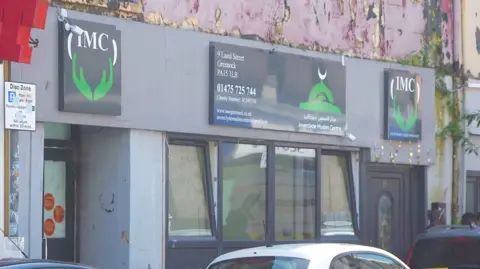 Exterior of a grey building with black doors and windows and peeling paint on the first storey. Above the entrance is a sign saying "IMC" for Inverclyde Muslim Centre.
