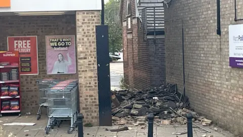 Broken tiles and bricks are in a pile in an alleyway. Shopping trolleys and a stand with coal bags are outside a store. 