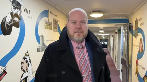 Shaun Whitmore/BBC John Neenan is wearing a thick coat with the collar turned up and a suit with a red striped shirt and tie. He is standing in a school corridor painted with murals.