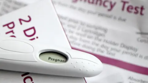 A positive pregnancy test lies on top of the test box which is white with magenta writing, the instructions for the kit are blurred out in the background.