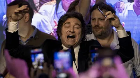 Getty Images Javier Milei vestindo terno, cercado de pessoas e com smartphones em primeiro plano filmando-o, joga as mãos para o alto com a boca aberta