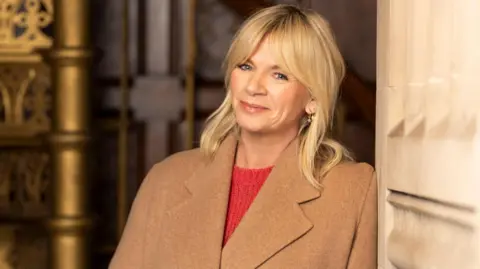 BBC The image shows a woman wearing a red jumper and brown coat, leaning against a wall.