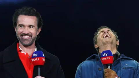 Rob Mac and Ryan Reynolds holding microphones and laughing as they stand against a dark backdrop. Rob wears a red shirt with a black coat, while Ryan wears a blue jacket and brown gloves