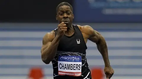Martin Rickett/PA Wire Dwain Chambers runs in the 60m during day one of the 2024 Microplus UK Athletics Indoor Championships at the Utilita Arena, Birmingham. He is wearing a black running vest with a sign attached to the front of it which reads CHAMBERS. He is grimacing as he exerts maximum effort. 