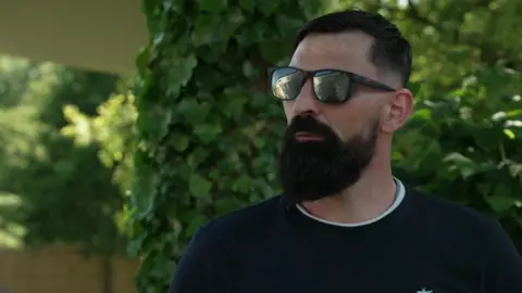 Dan Stanaway wears reflective sunglasses as he stands in front of a hedge outside his old boarding school. He has short dark hair and a long dark beard.