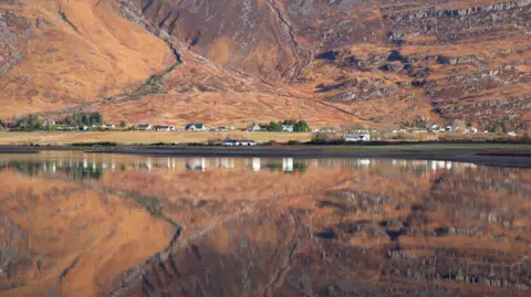 Torridon village and the mountainside that rises behind it. The village and mountain are reflected in the mirror-like waters of Loch Torridon.