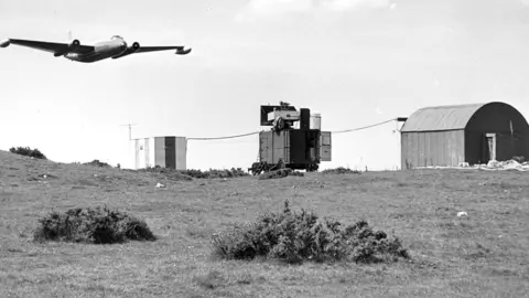 A black and white photograph showing a plane flying overhead as a mobile radar tracks it