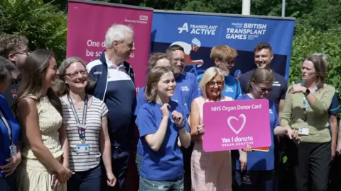 A group of people standing together, whilst one in the middle holds a large pink organ donation card.