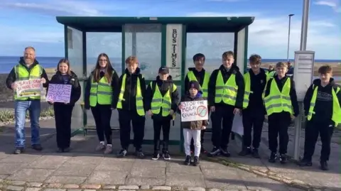 Campaigners with save school bus signs at a bus stop, wearing yellow hi-vis tops.