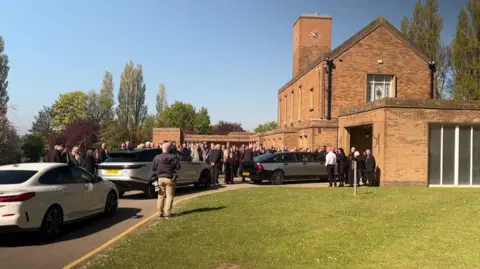 A crematorium building with dozens of people stood outside. There is black car and two white cars approaching the building.