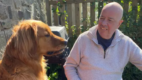 A man wearing a light-coloured jumper is looking at a dog who is sat to the side of him. They are outdoors, in what appears to be a garden with a fence nearby behind them.