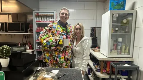 Bournville Cafe A man and woman are standing behind a cafe counter together. The woman has bobbed blonde hair and is wearing a cream tracksuit.The man has short blonde hair and is wearing a jacket filled with rubber ducks and stuffed teddies. He is also holding a white mug. They have their arms around each other and are stood smiling at the camera.