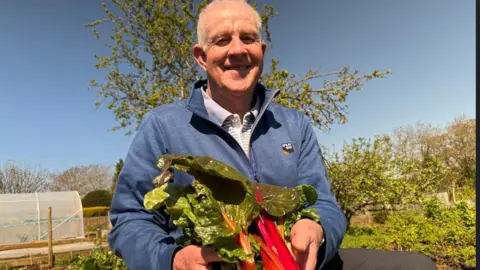 Chris McHugh / BBC A man poses in his allotment holding a bunch of freshly cut rainbow chard, which has large green leaves and pink branches.