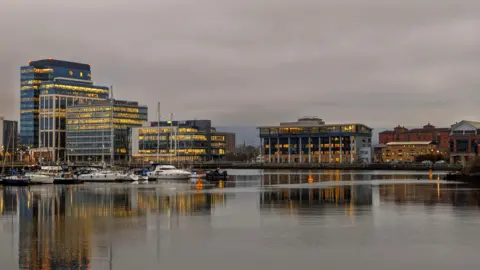 BBC Weather Watcher A-Lo Belfast view from the Maritime Mile showcasing boats docked in the harbour and buildings lit up in the early evening. The sky in the background has a pink tone to it but clouds fill the sky. 