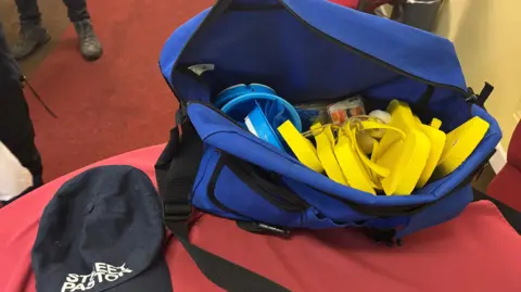 A navy cap with white writing on it that says "stafford street pastors". there is a blue bag containing yellow flip flops and blue plastic bags 