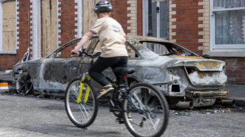 A burnt out car is on a street. Boarded up terraced houses are behind it. There is a kid on a bike in front of the burnt-out car.