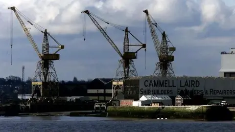 PA Media A general view of the Cammell Laird shipyard in the river Mersey in Birkenhead, Merseyside. The image shows three large cranes and the wall of a warehouse on which the words Cammell Laird Shipbuilders, Engineers and Repairers is written in large capital letters. 