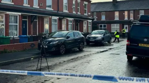 BBC Police tape across a residential street in Moss Side. There is a camera on a tripod in the road, and two police officers in uniform in the backdrop. A bicycle is on its side next to a car within the cordon. 