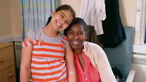 Family Jasmine when she was young. She is wearing a grey and orange striped top and is standing next to her mother, who is sitting in a chair. The pair seem to be on a hospital ward or in a side room - there is a radiator behind them and a cabinet to the left