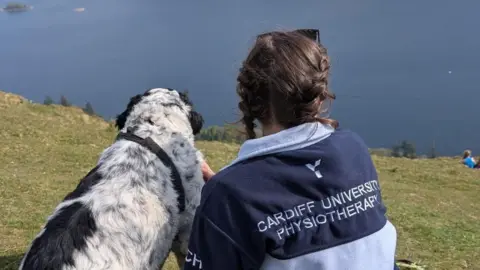 Chloe Hurst A woman and dog look out at a lake on a sunny day. The dog on the left is large and has black and white fur. On the right, Chloe wears a navy and blue Cardiff University physiotherapy fleece and has her hair in french plaits.