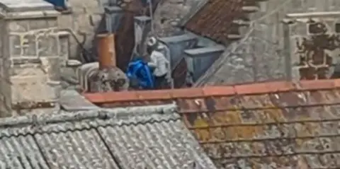 Avon & Somerset Police CCTV image of a man on the roof of a shopping centre. The image is very blurry. He can be seen carrying a large, dark blue item. 