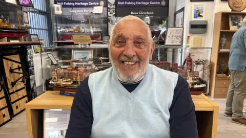 Terence Thresh sits in front of model ships in the Hull Fishing Heritage Centre. He looks directly at the camera with a big smile, wearing a dark blue long sleeved top and light blue vest. 