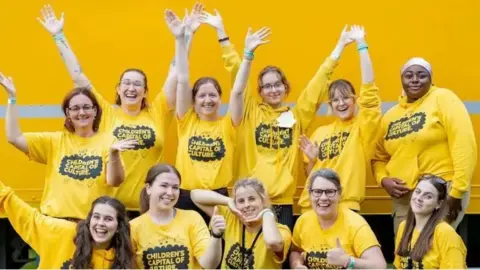 A group of 11 young women, in two rows, wave their arms or give thumbs up in front of a yellow background. They are wearing yellow shirts and hoodies, all emblazoned with a "Children's Capital of Culture" logo.