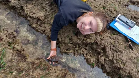 A woman smiling at the camera while lying on a rock. Her hand is in a rockpool holding some kelp leaves.