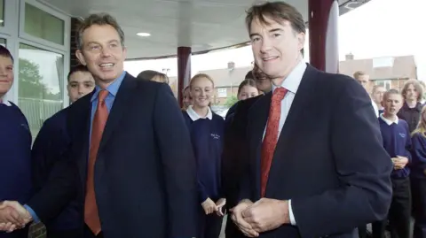 PA Media Then-Prime Minister Tony Blair with Peter Mandelson in 2001. They are walking past a group of pupils on a visit to a school in Hartlepool.