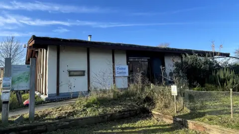 FOLKESTONE AND HYTHE DISTRICT COUNCIL The Romney Marsh Visitor Centre, with white walls and a dark brown roof and wall beams, with vegetation in front of the building and a blue sky behind it.