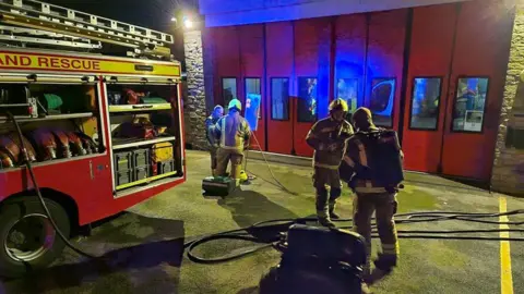 On-call firefighters outside a fire station at night, next to an engine and hose.