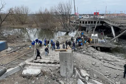 Orla Guerin/BBC People fleeing Irpin cross the damaged river bridge, which can be seen with the concrete collapsed and girders hanging down into the water