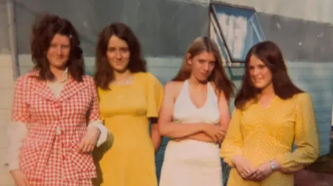 Susan Morris The same four women against the backdrop of a caravan in 1972. Two of them are wearing bright yellow dresses, another white, and the other a red and-white chequered top and skirt.